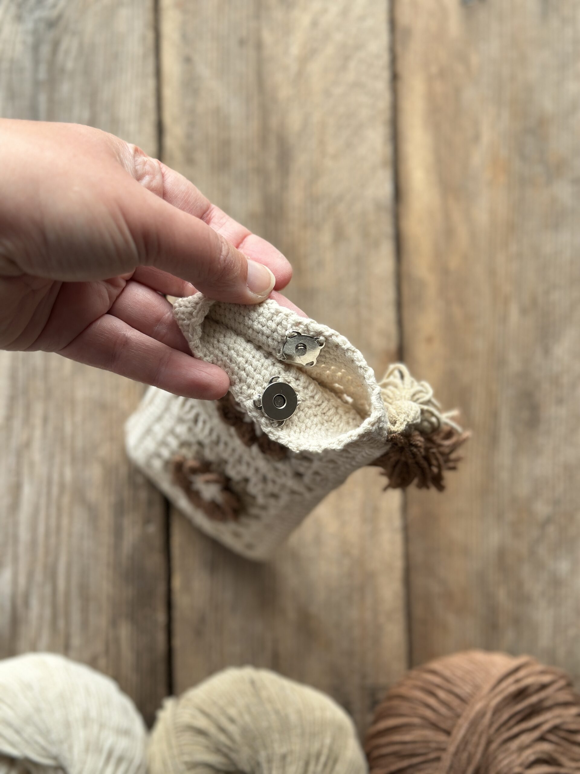szydełkowe etui na telefon lub okulary, zapinane na zapięcie magnetyczne, ozdobione dwoma chwostami. kolor dominujący to jasny beż, dodatkowo brąz i ciemny beż.