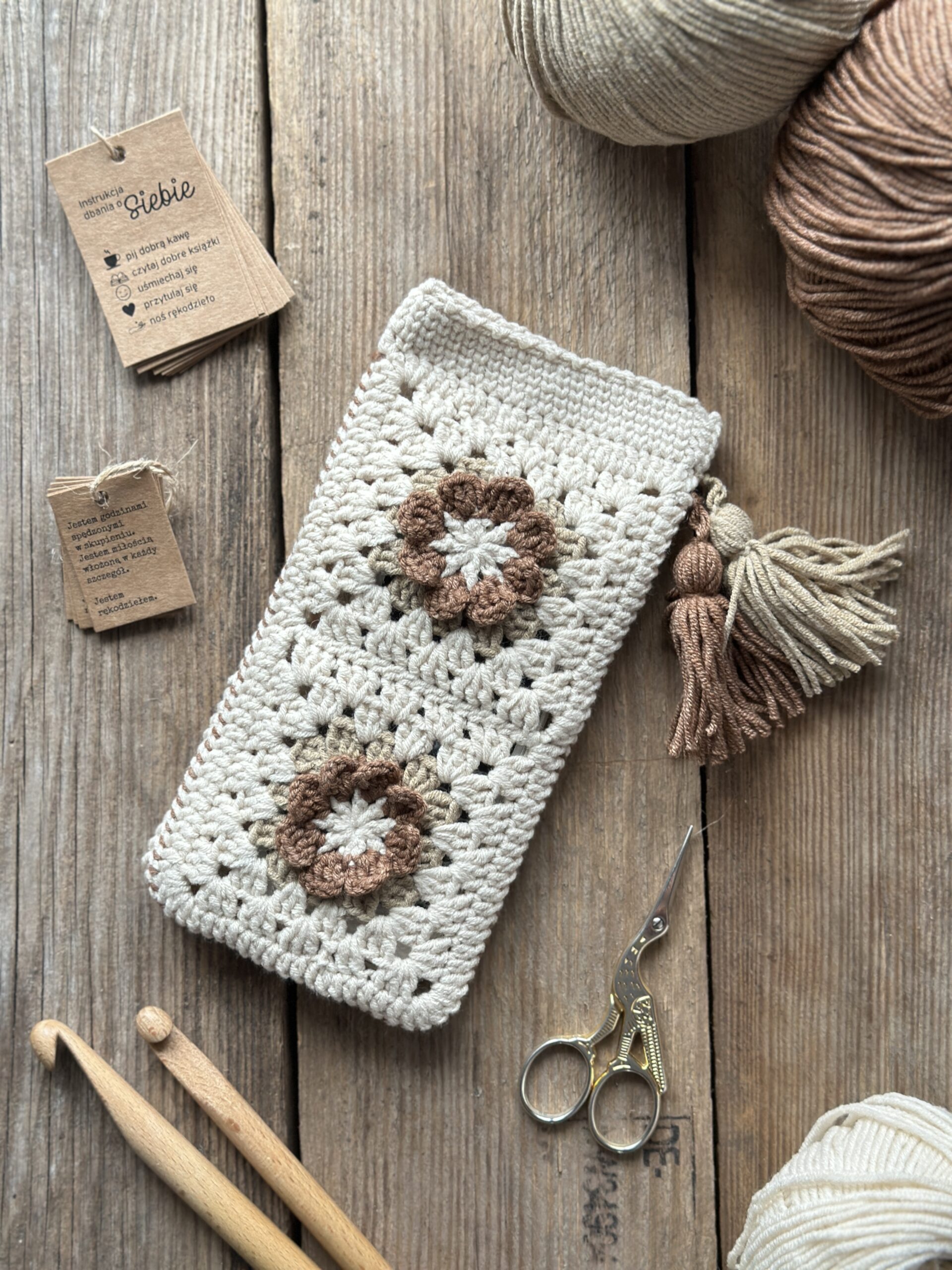 szydełkowe etui na telefon lub okulary, zapinane na zapięcie magnetyczne, ozdobione dwoma chwostami. kolor dominujący to jasny beż, dodatkowo brąz i ciemny beż.