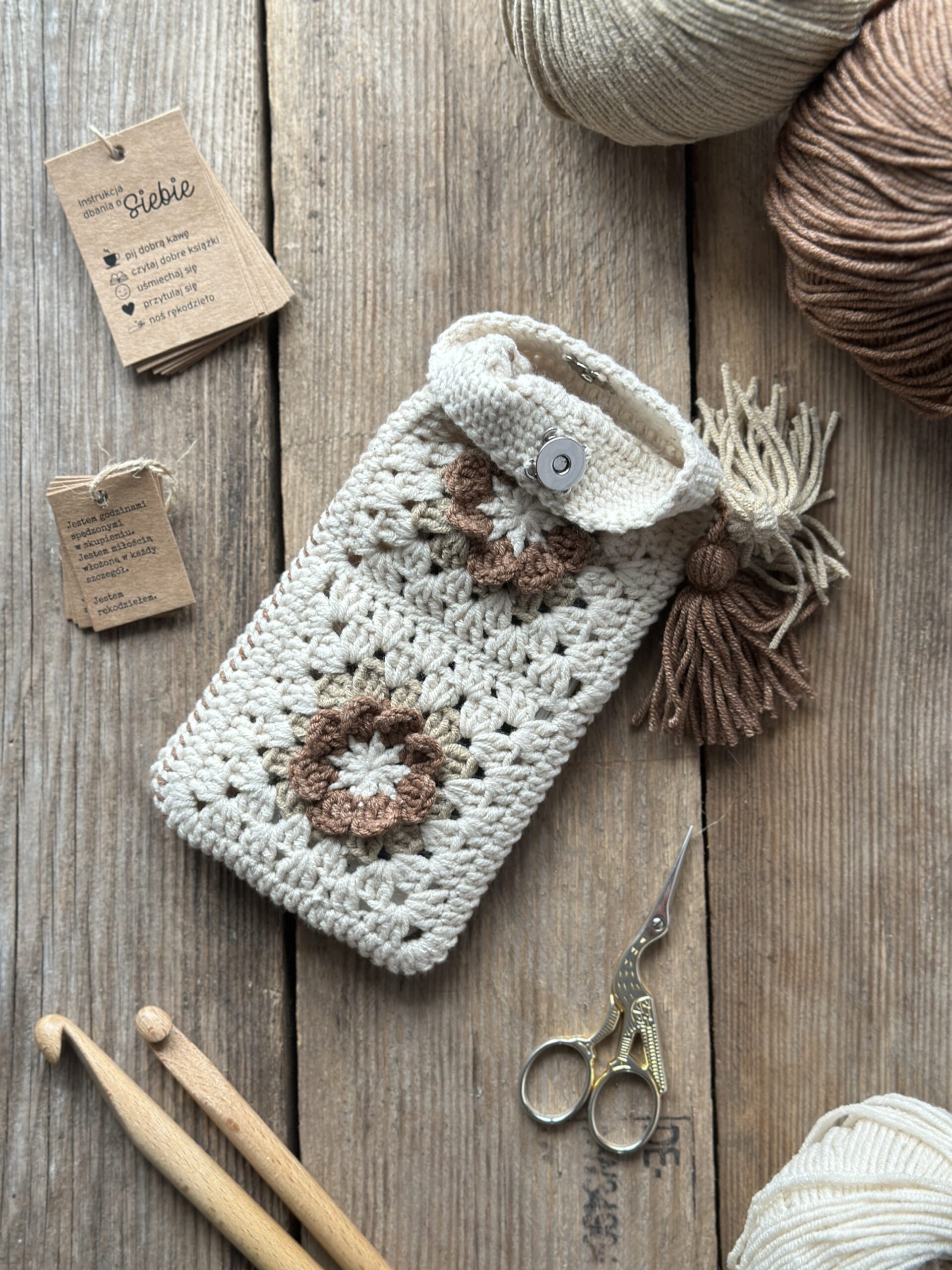 szydełkowe etui na telefon lub okulary, zapinane na zapięcie magnetyczne, ozdobione dwoma chwostami. kolor dominujący to jasny beż, dodatkowo brąz i ciemny beż.